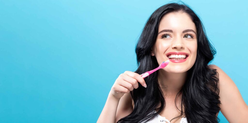 水色の背景の前でピンクの歯ブラシを片手に持つ笑顔の女性